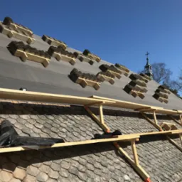 Montaż nowej dachówki karpiówki na dachu z widocznym fragmentem starego pokrycia, rusztowaniem i sylwetką kościoła w tle.