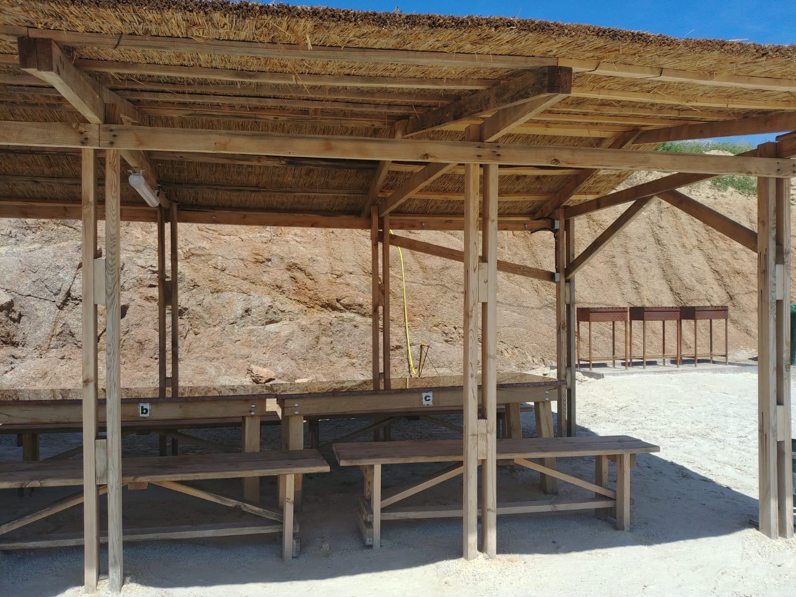 Zadaszona drewniana konstrukcja z trzcinowym dachem, stoliki z oznaczeniami literowymi na piaskowym podłożu, skaliste tło.