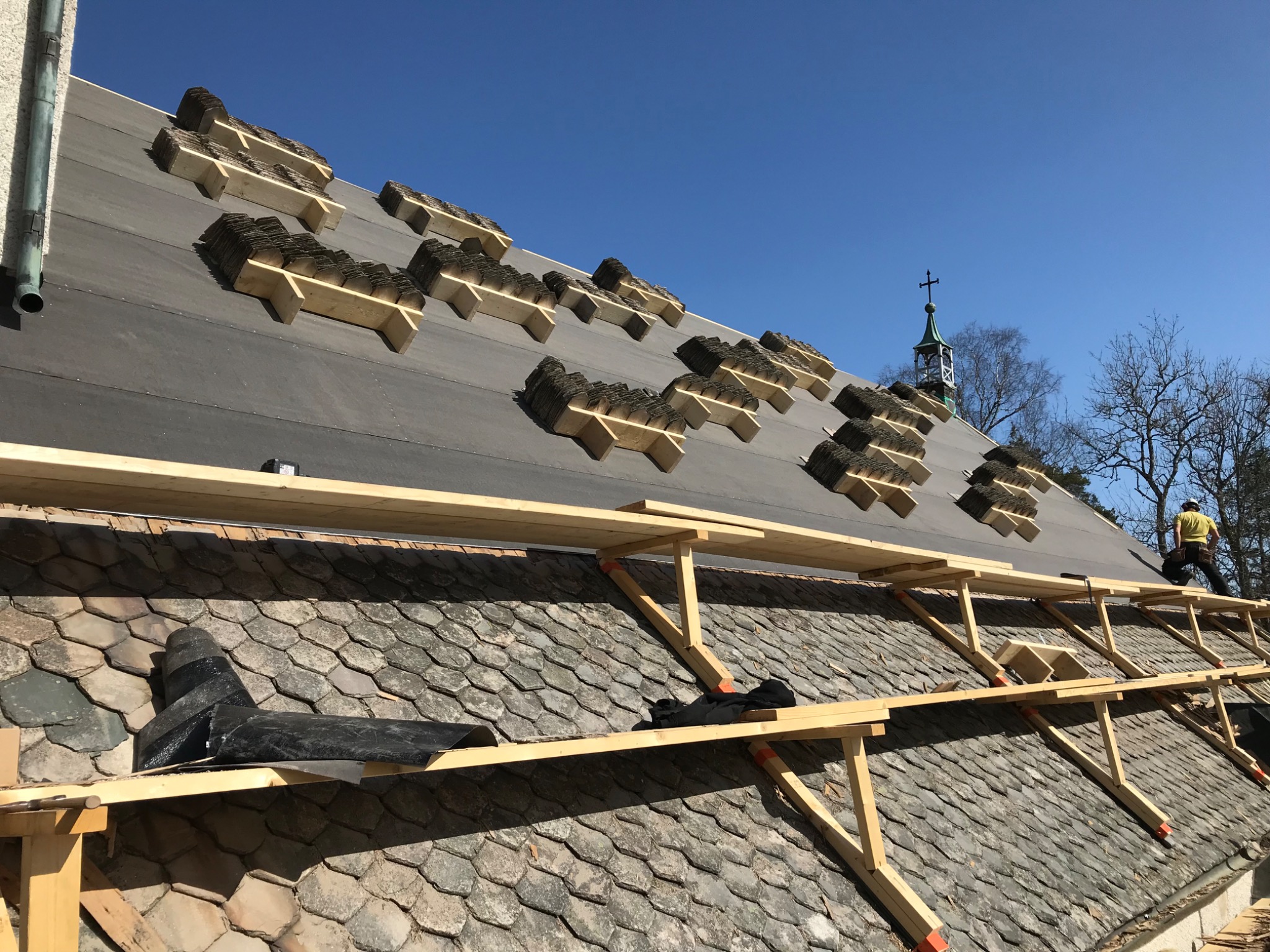 Montaż nowej dachówki karpiówki na dachu z widocznym fragmentem starego pokrycia, rusztowaniem i sylwetką kościoła w tle.
