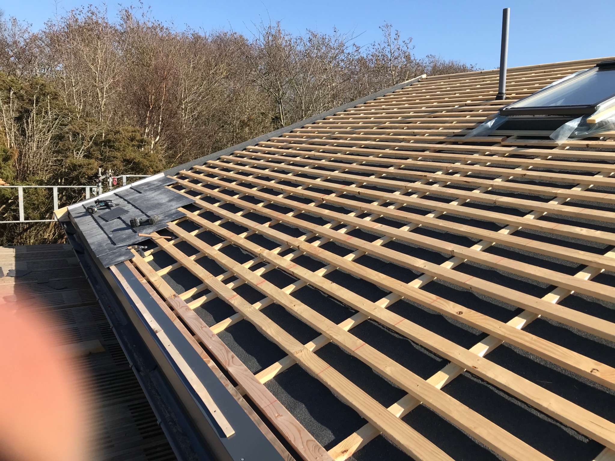 Montaż pokrycia dachowego z łupka na drewnianej konstrukcji w Legnicy, widoczne okno dachowe i komin na tle drzew bez liści.