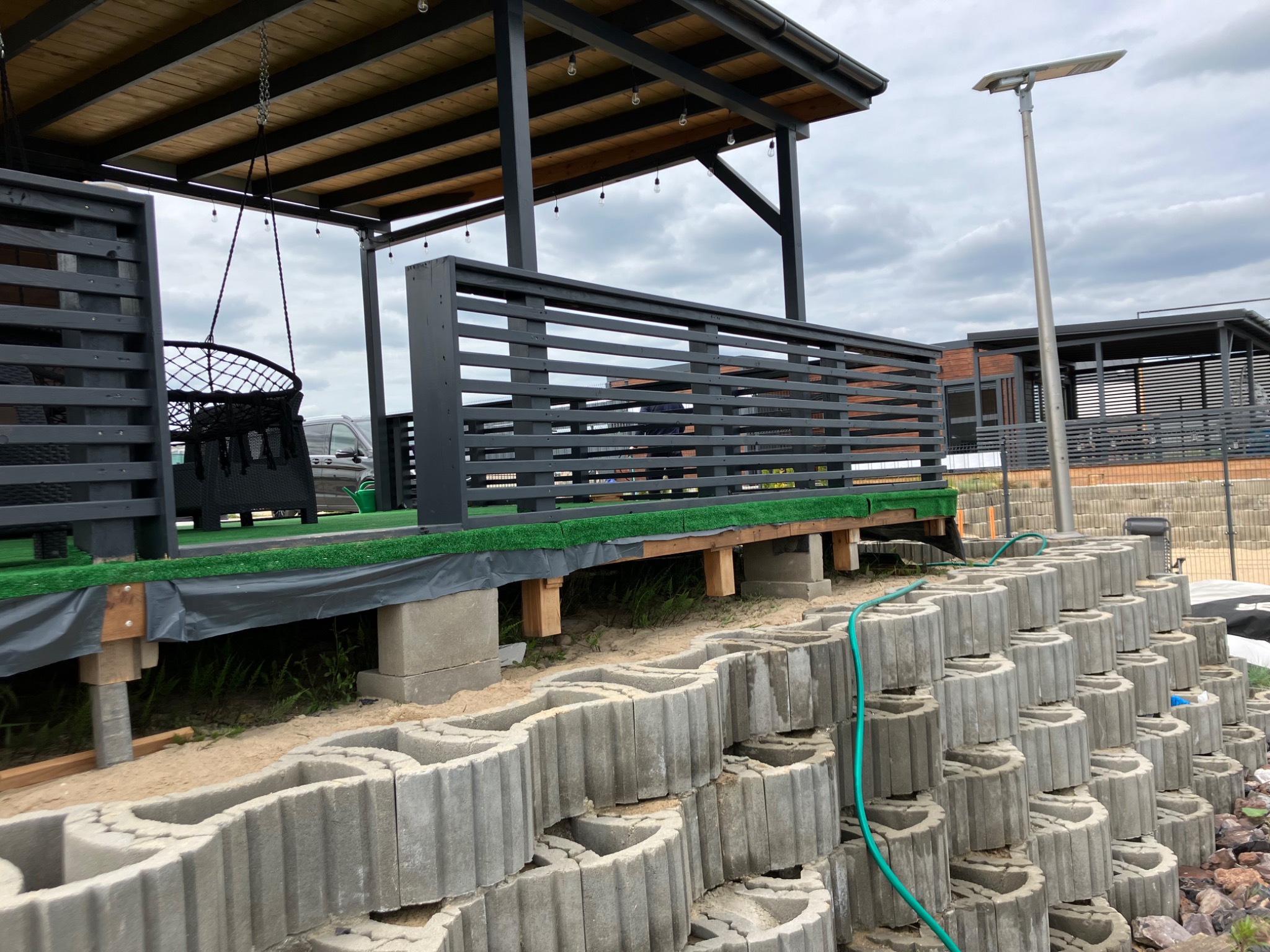 Budowa tarasu z szarej, drewnianej konstrukcji i betonowych bloczków na nierównym terenie, widoczny fragment ogrodu i oświetlenie zewnętrzne.