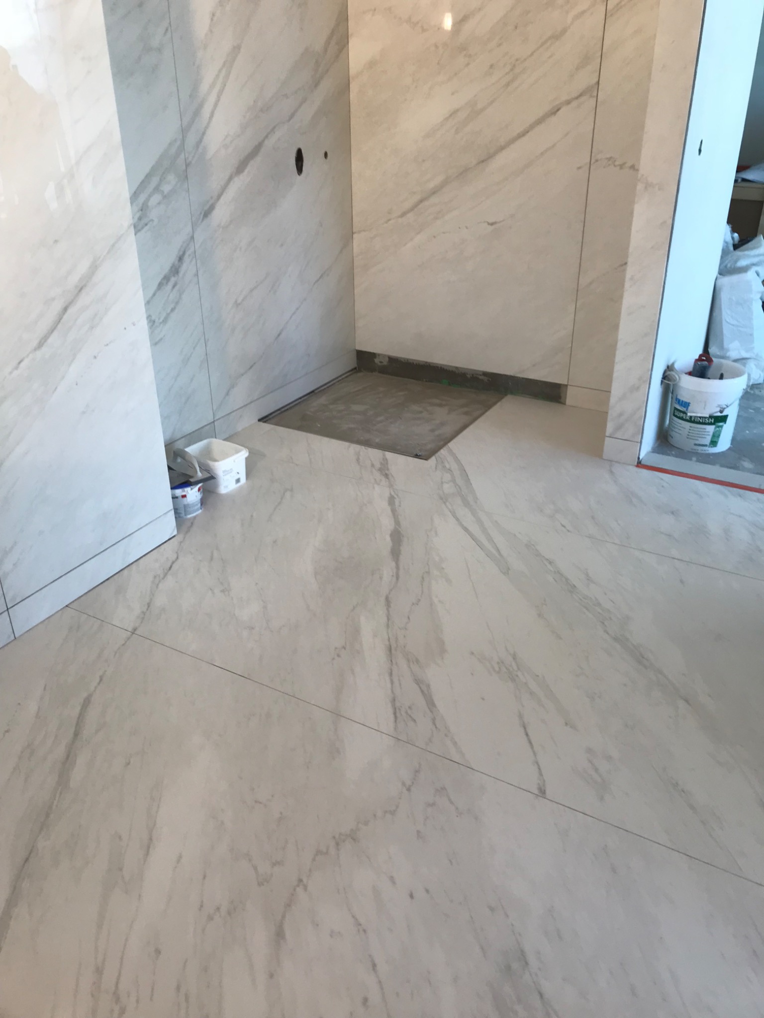 Wnętrze łazienki w trakcie prac wykończeniowych, widoczne płytki imitujące marmur na ścianach i podłodze, przygotowane miejsce na brodzik, wiaderka z zaprawą i narzędzia.