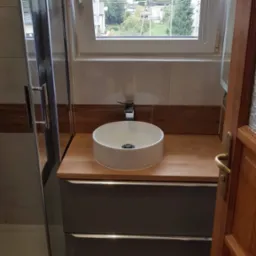 Nowoczesna łazienka z okrągłą umywalką nablatową, drewnianym blatem, szarą szafką pod umywalką i częściowo widoczną kabiną prysznicową z przezroczystego szkła, widok zza uchylonych drewnianych drzwi.