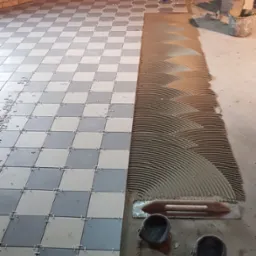 Układanie szachownicy z szarych i kremowych płytek podłogowych, z krzyżykami dystansowymi, widoczny klej na podłodze, kielnia i dwa małe wiadra na zaprawę.