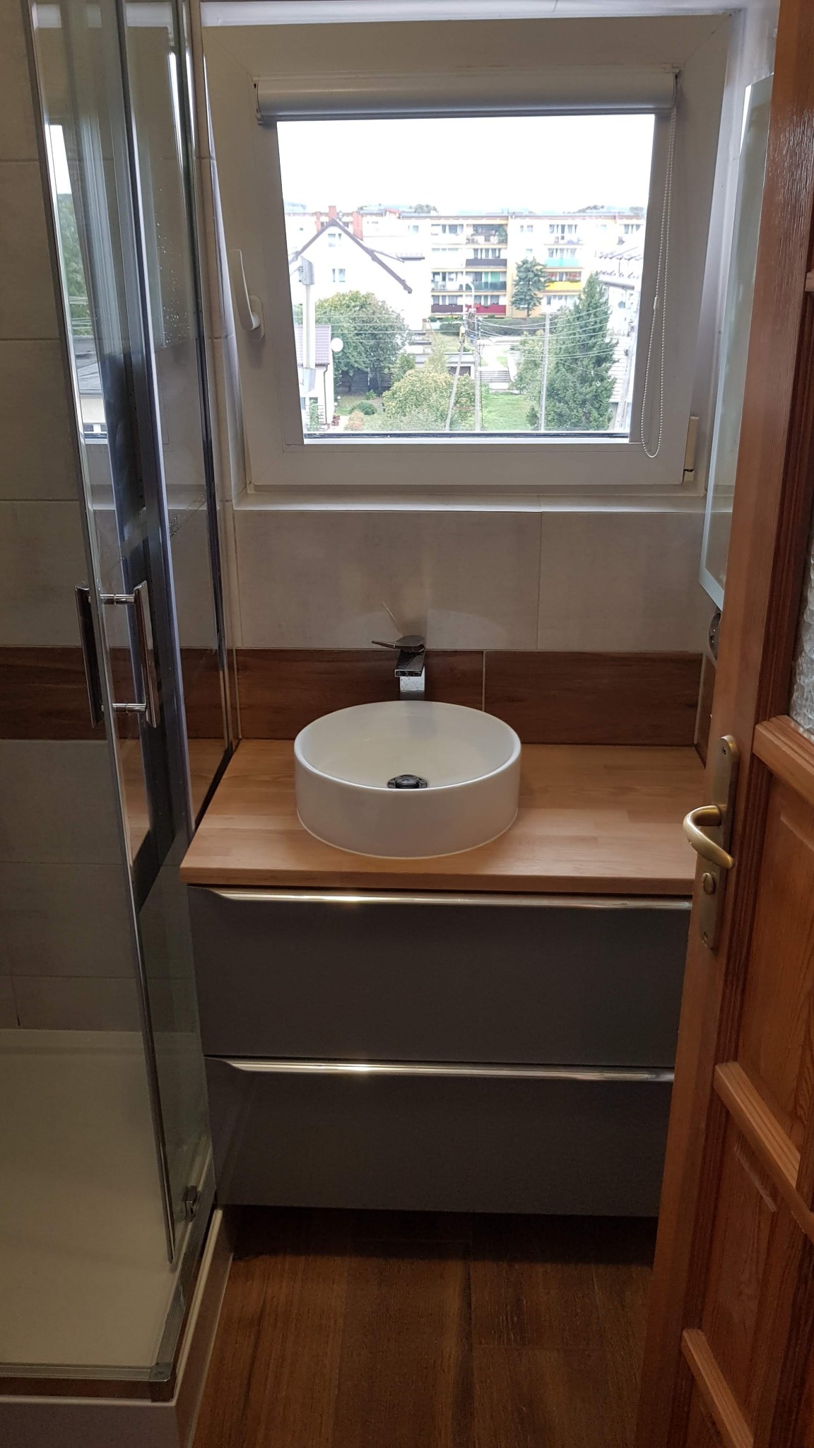 Nowoczesna łazienka z okrągłą umywalką nablatową, drewnianym blatem, szarą szafką pod umywalką i częściowo widoczną kabiną prysznicową z przezroczystego szkła, widok zza uchylonych drewnianych drzwi.