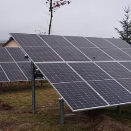 Dwa rzędy paneli słonecznych na metalowych konstrukcjach, zainstalowane na trawiastym terenie z drzewami w tle, widoczne ślady po instalacji.