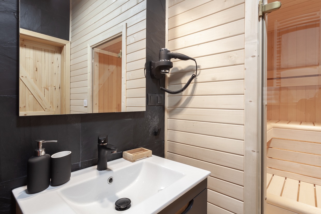 Nowoczesna łazienka z elementami drewna: biała umywalka z czarną armaturą, czarne akcesoria łazienkowe, lustro odbijające drewniane drzwi, suszarka do włosów na ścianie i widoczna sauna...