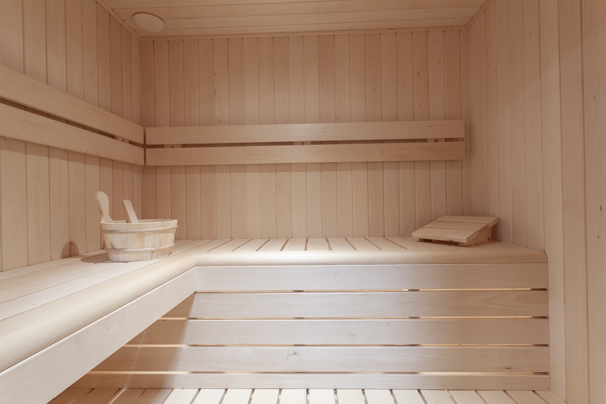 Jasne, drewniane wnętrze sauny z ławkami, oświetleniem sufitowym i akcesoriami: wiaderkiem z chochlą oraz zagłówkiem.