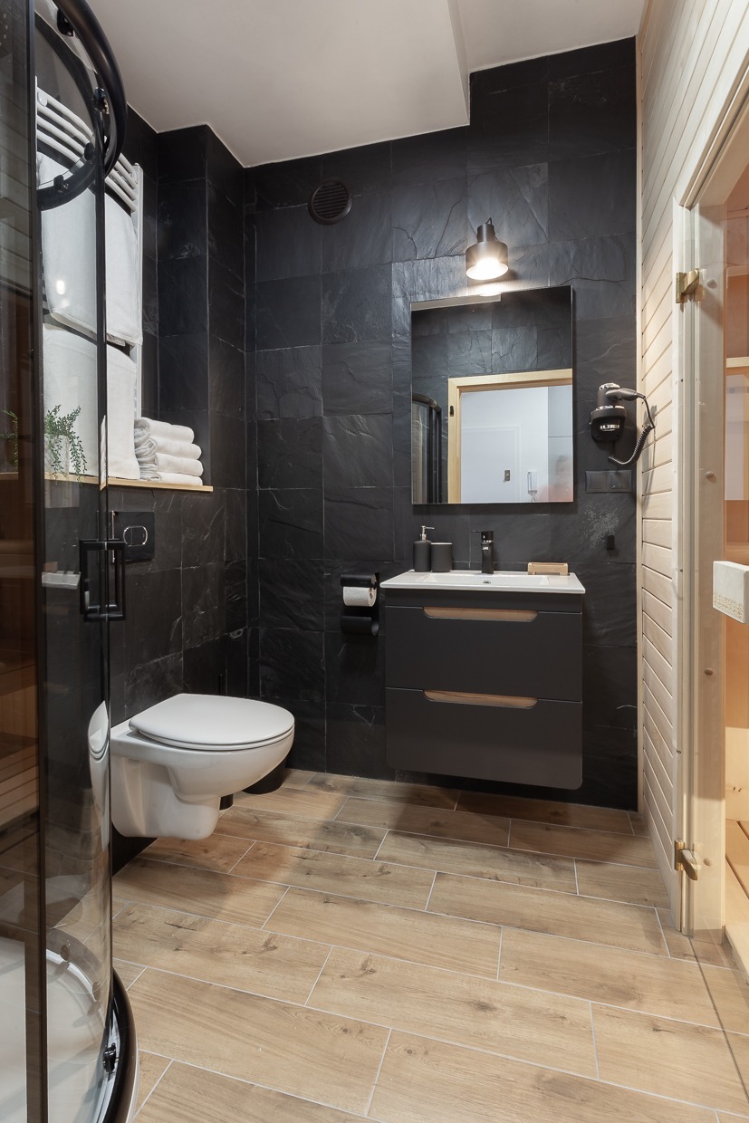Nowoczesna łazienka z czarnymi płytkami imitującymi kamień, drewnopodobną podłogą, wiszącą toaletą, szarą szafką pod umywalką i kabiną prysznicową z zaokrąglonym szkłem.