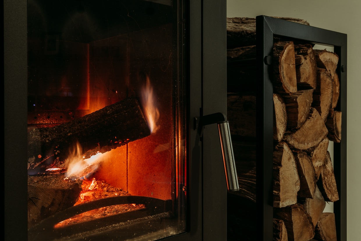 Płonące drewno w nowoczesnym, czarnym kominku z szybą, obok stojak na drewno.
