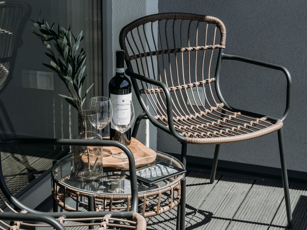 Rattanowe krzesła i stolik z butelką wina na balkonie z drewnianą podłogą i szarą ścianą, w tle widoczna przeszklona ściana z zasłoną.
