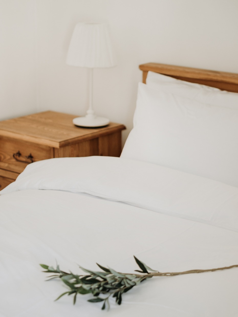 Jasna sypialnia z drewnianym łóżkiem, białą pościelą i gałązką oliwną na kołdrze, obok drewniana szafka nocna z białą lampką.