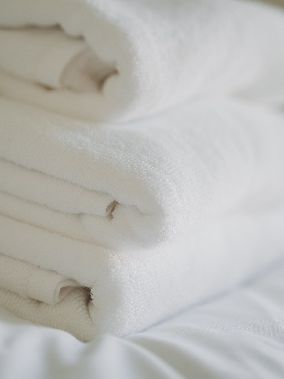 Stos białych, puszystych ręczników frotte ułożonych na białej pościeli, z delikatnym cieniem podkreślającym fakturę.