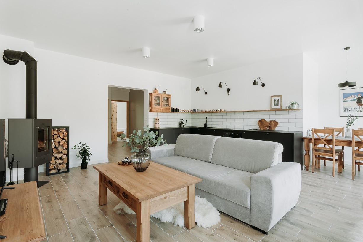 Jasne wnętrze z drewnianą podłogą, piecem na drewno z zapasem drewna, szarą sofą i drewnianym stolikiem kawowym z dekoracyjnym wazonem, w tle czarna zabudowa kuchni z białymi płytkami.