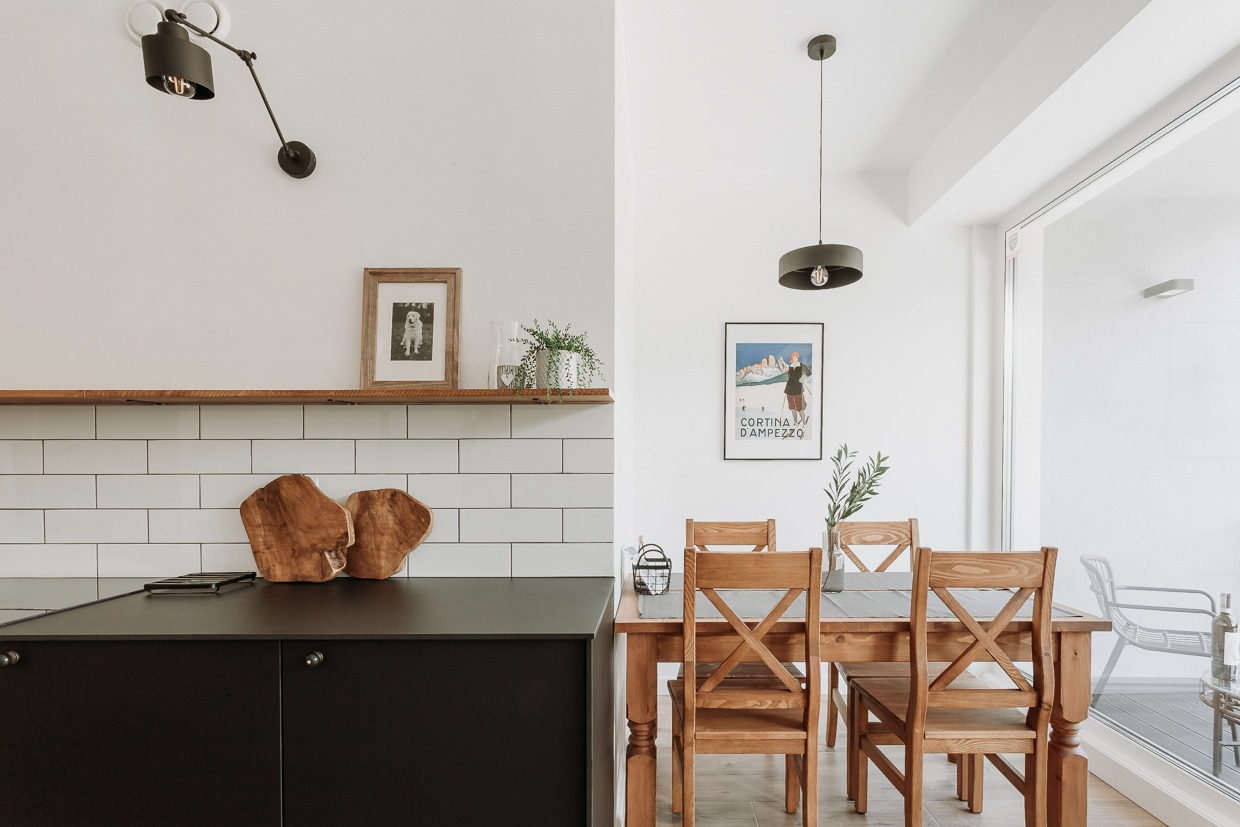 Jasne wnętrze: drewniany stół z krzesłami, czarne szafki kuchenne, białe płytki, drewniana półka z dekoracjami, plakat na ścianie i nowoczesne oświetlenie.