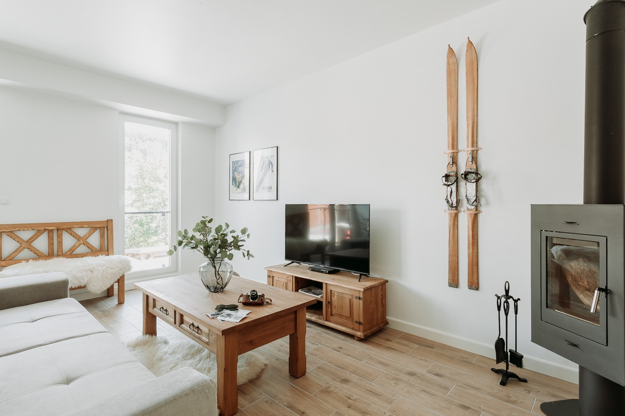 Jasne wnętrze salonu z drewnianymi elementami: stół z wazonem i zielonymi gałązkami, szafka pod telewizor, dekoracyjne stare narty na ścianie, nowoczesny kominek i drewniane łóżko z futrzanym pledem.