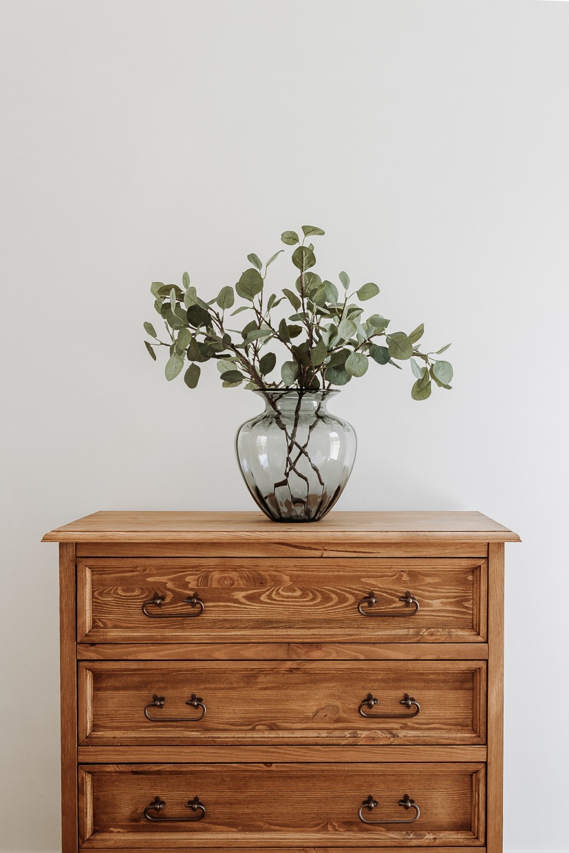 Drewniana komoda z trzema szufladami, na której stoi szary, szklany wazon z gałązkami eukaliptusa na tle białej ściany.
