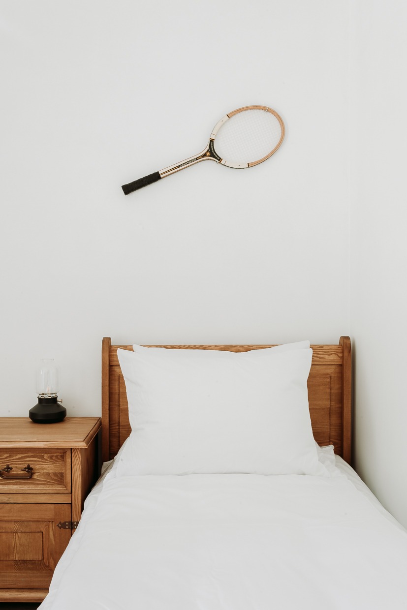 Pokój z łóżkiem pojedynczym, drewnianym stolikiem nocnym z lampką naftową i rakietą tenisową zawieszoną na ścianie.