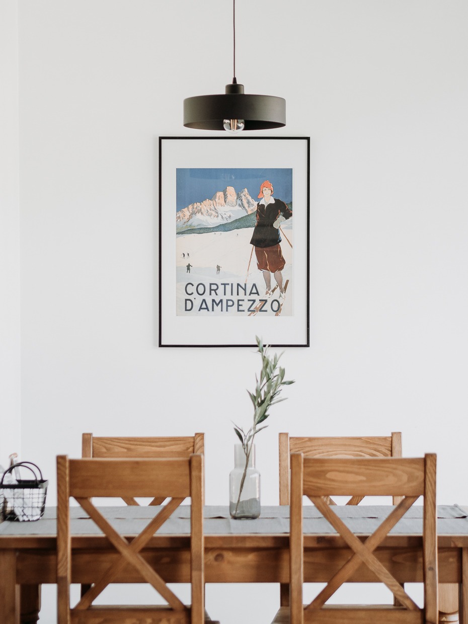 Jadalnia z drewnianym stołem i krzesłami typu X, nad stołem wazon z gałązką oliwną, na ścianie plakat w czarnej ramie z motywem górskim Cortina D'Ampezzo, oświetlenie stanowi czarna lampa sufitowa.