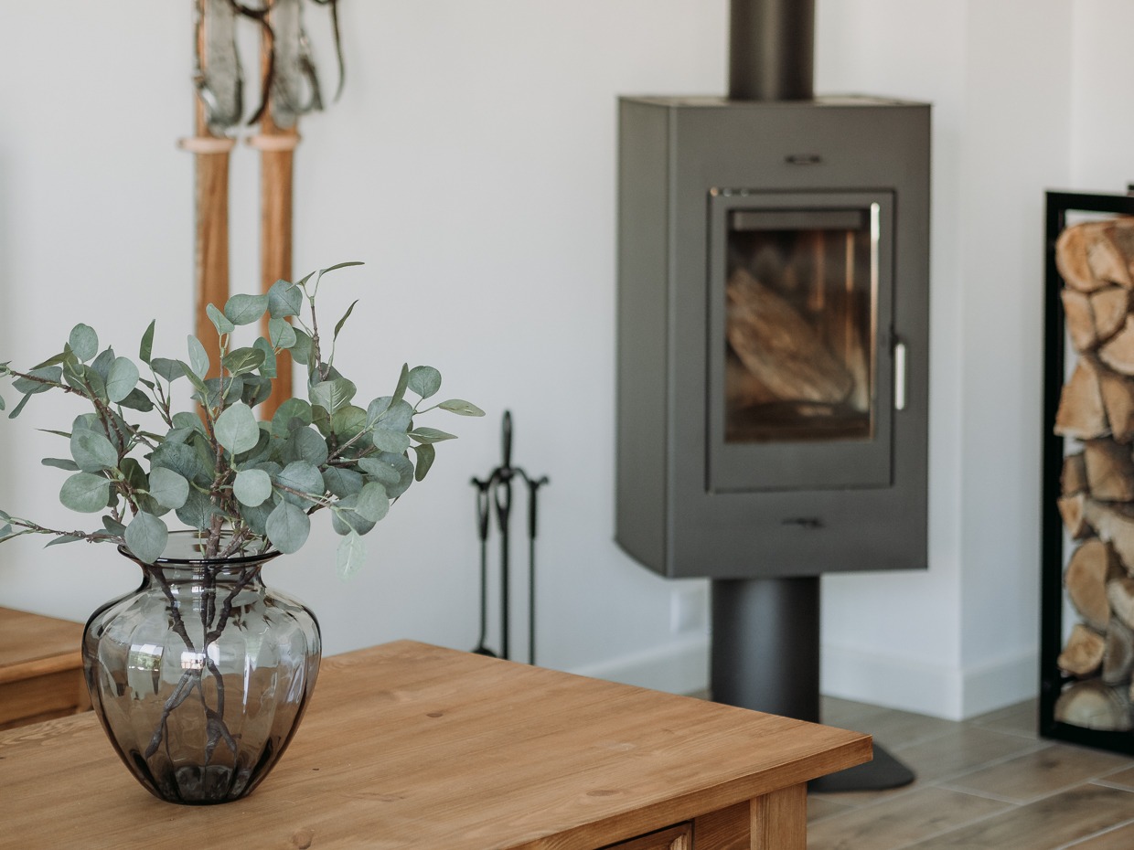 Nowoczesny salon z kominkiem wolnostojącym, drewnianym stolikiem i dekoracyjnym wazonem z eukaliptusem na blacie. W tle widoczne drewno kominkowe ułożone w czarnej ramie.