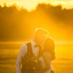 Szymon Gołasz - Zakochana para w objęciach, pocałunek na tle zachodzącego słońca, on w kamizelce i muszce, ona w białej sukience trzyma bukiet kwiatów.