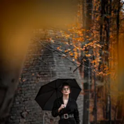 Portret kobiety w okularach, trzymającej czarną parasolkę, ubrana w czarny płaszcz z ozdobnym paskiem, na tle jesiennego lasu i ceglanej ruiny.