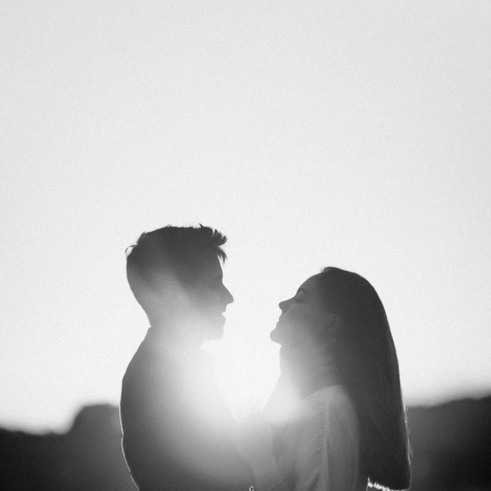 Czarno-biała fotografia sylwetek pary zakochanych, zwróconych do siebie twarzami na tle zachodzącego słońca, tworzącego efekt aureoli.