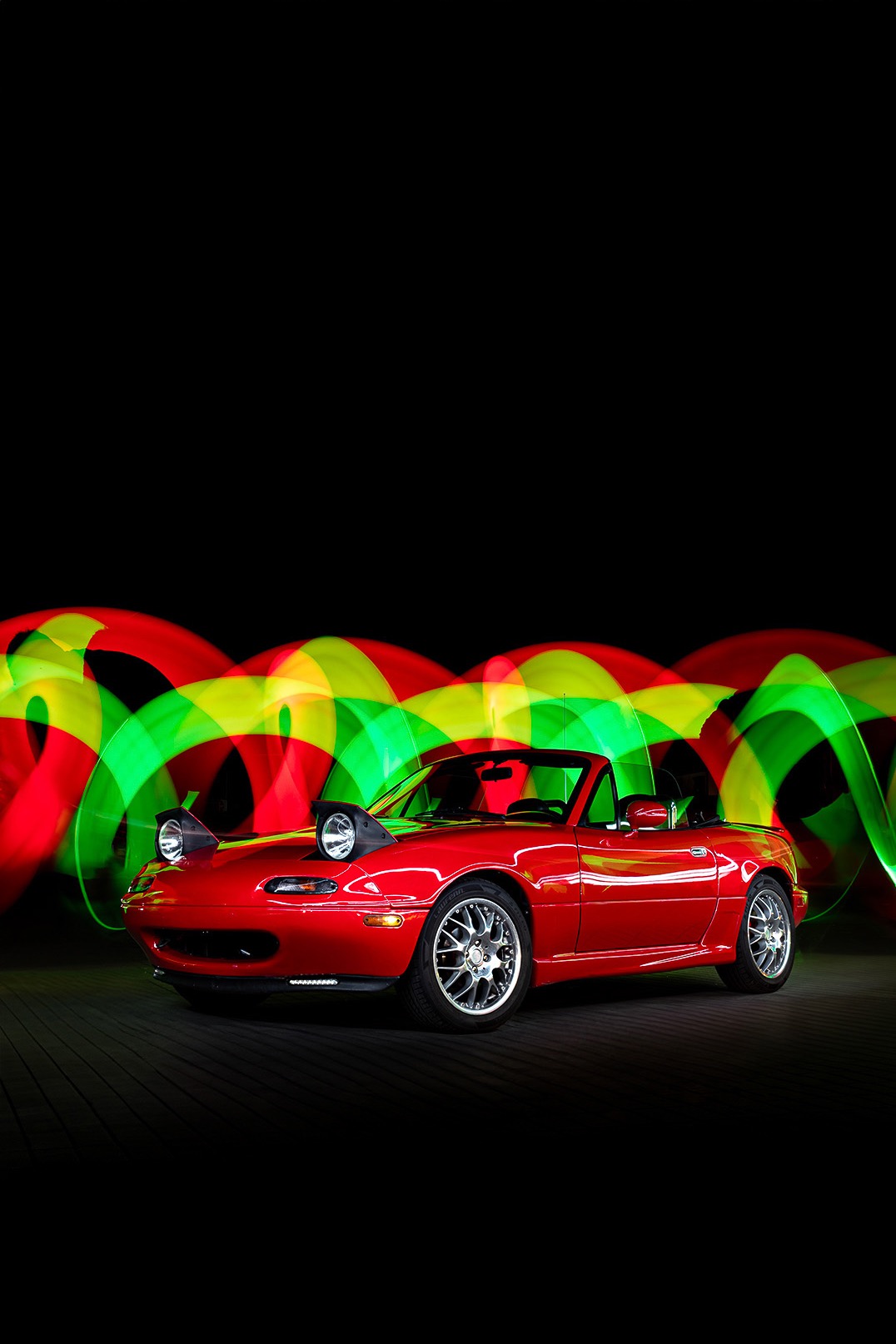 Czerwony kabriolet Mazda Miata z podniesionymi reflektorami na tle dynamicznych smug światła w kolorach czerwonym, żółtym i zielonym, tworzących efektowną kompozycję.