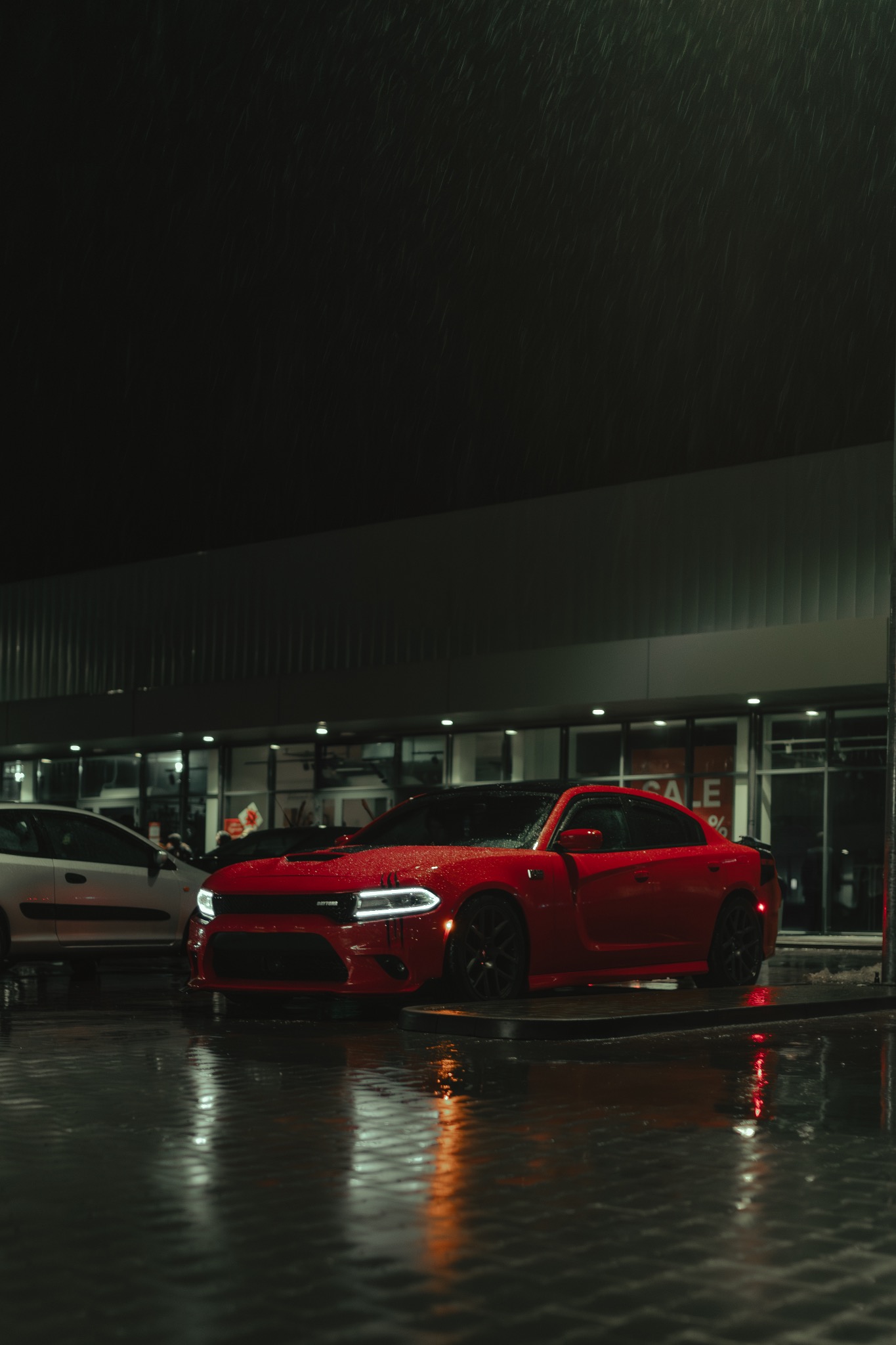 Czerwony Dodge Charger Daytona zaparkowany w deszczową noc przed budynkiem z oknami wystawowymi, widoczne krople deszczu na karoserii i mokry asfalt.