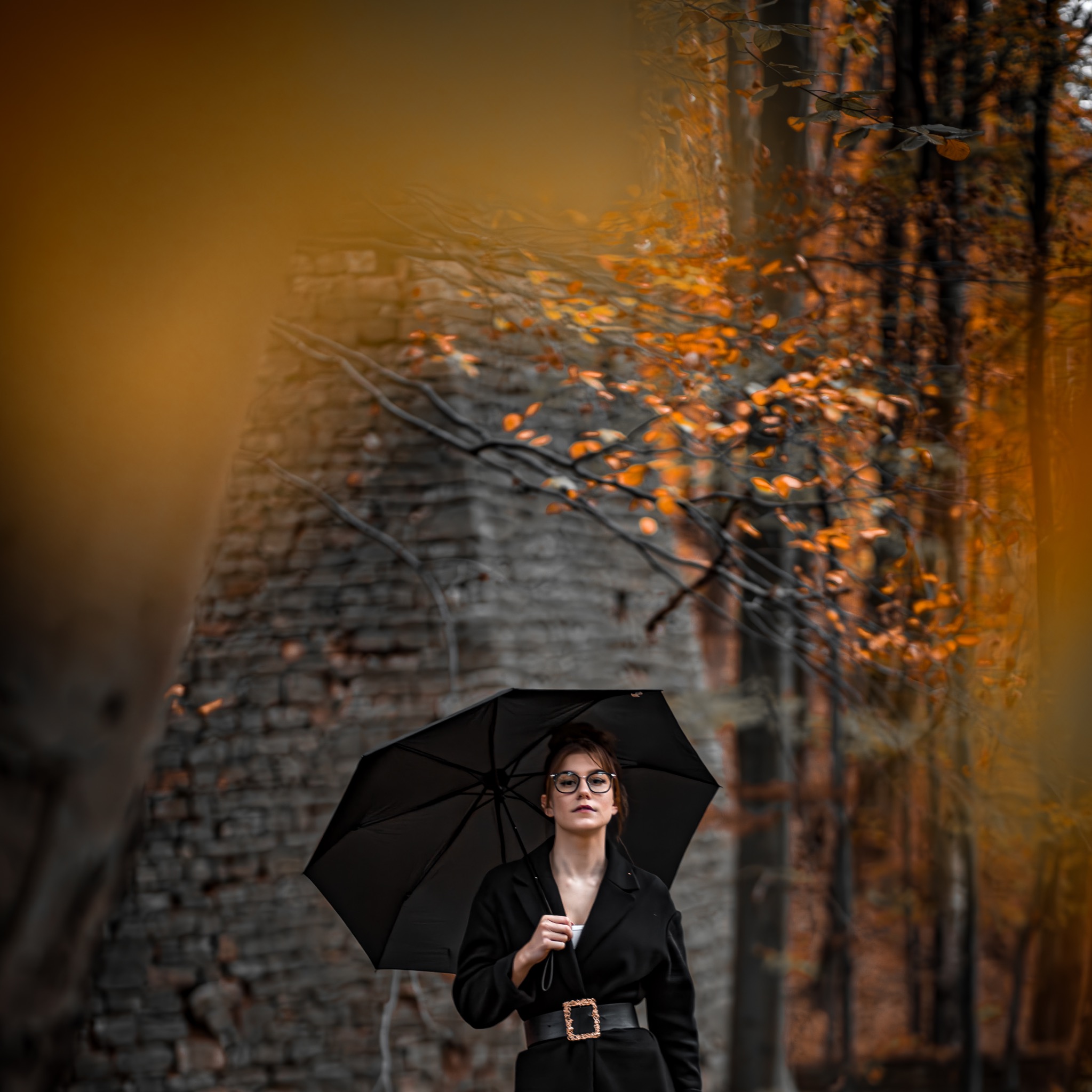 Portret kobiety w okularach, trzymającej czarną parasolkę, ubrana w czarny płaszcz z ozdobnym paskiem, na tle jesiennego lasu i ceglanej ruiny.