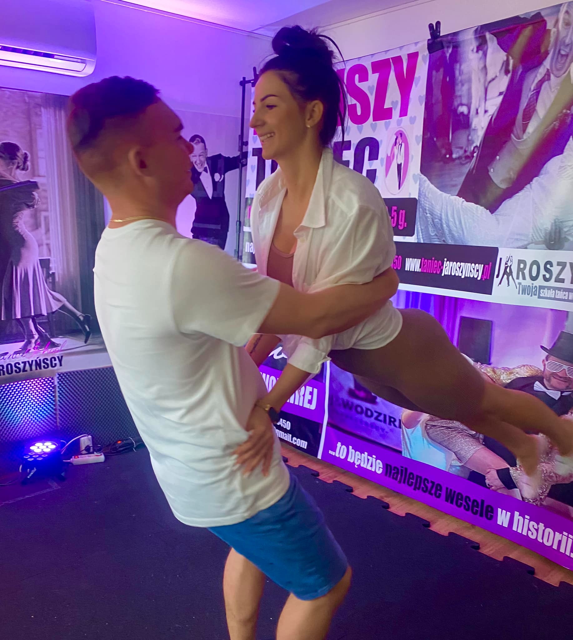 Para taneczna w trakcie dynamicznego podnoszenia na tle plakatu reklamowego z hasłem 'to będzie najlepsze wesele w historii'. Kobieta w białej koszuli i legginsach uśmiecha się, unoszona przez...