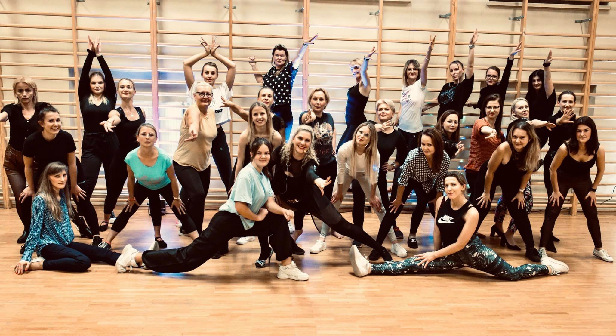 Grupa kobiet w różnym wieku pozuje w sali gimnastycznej, wykonując ruchy taneczne, niektóre siedzą na podłodze, inne stoją, w tle drabinki gimnastyczne.