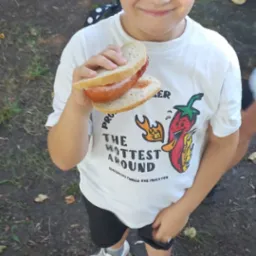 Uśmiechnięty chłopiec z kanapką z kiełbasą w dłoni, ubrany w białą koszulkę z motywem papryczki chili i czarne spodenki, stojący na ziemi pokrytej liśćmi.