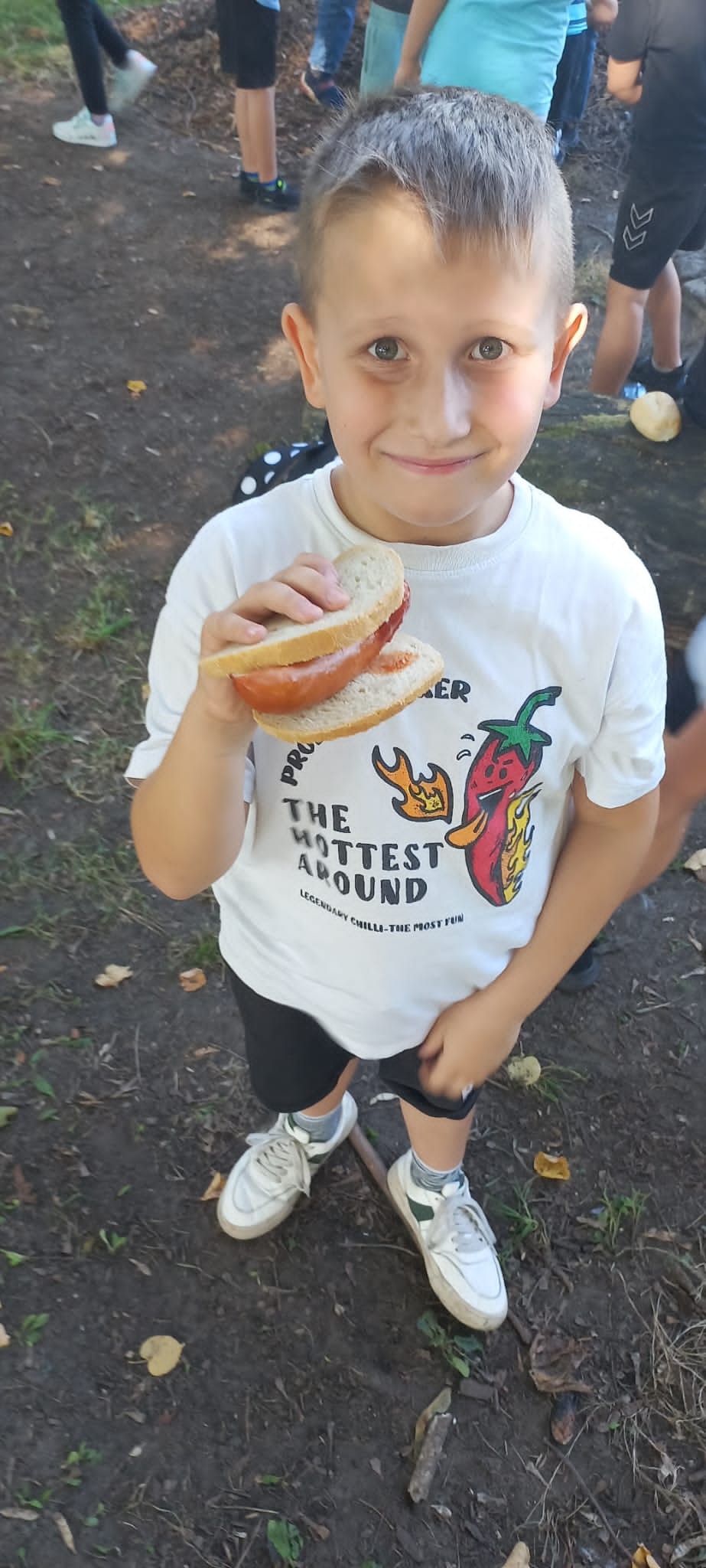 Uśmiechnięty chłopiec z kanapką z kiełbasą w dłoni, ubrany w białą koszulkę z motywem papryczki chili i czarne spodenki, stojący na ziemi pokrytej liśćmi.