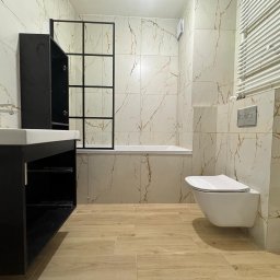 ANDRZEJ ŁYSAKOWSKI USŁUGI BUDOWLANO-TRANSPORTOWE - Wykończona łazienka w jasnych płytkach imitujących marmur z akcentami złota, z czarną szafką pod umywalką i kabiną prysznicową typu loft z czarnymi profilami, toaleta podwieszana.
