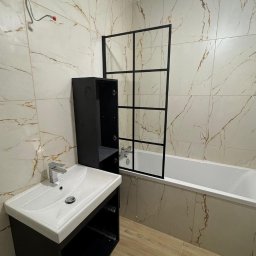 ANDRZEJ ŁYSAKOWSKI USŁUGI BUDOWLANO-TRANSPORTOWE - Nowoczesna łazienka z marmurowymi płytkami, czarną kabiną nawannową z geometrycznymi podziałami, białą umywalką z szafką w kolorze antracytu oraz drewnopodobną podłogą. Widoczne niedokończone...