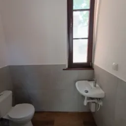 Jasna łazienka po remoncie: WC, umywalka pod oknem z brązową ramą, szare płytki do połowy ściany, drewnopodobna podłoga. Nowoczesne i funkcjonalne wykończenie.