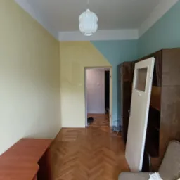 Pokój przed remontem: parkiet w jodełkę, ściany żółto-turkusowe z białym sufitem, wisząca lampa, meble w tle, drzwi oparta o szafę. Widok na wnętrze przez otwarte drzwi.