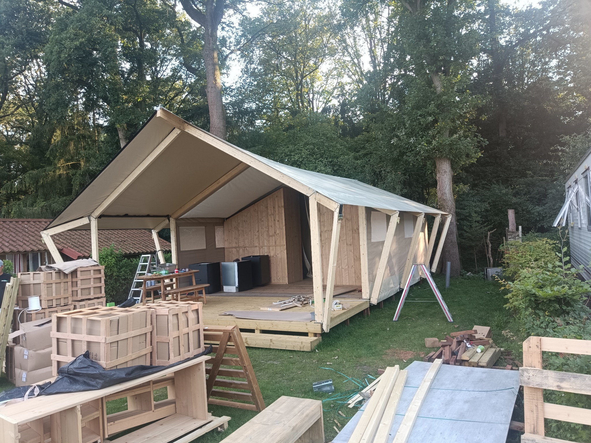 Budowa nietypowej drewnianej konstrukcji z elementami płótna na tle drzew, widoczne materiały budowlane i narzędzia, etap wznoszenia.