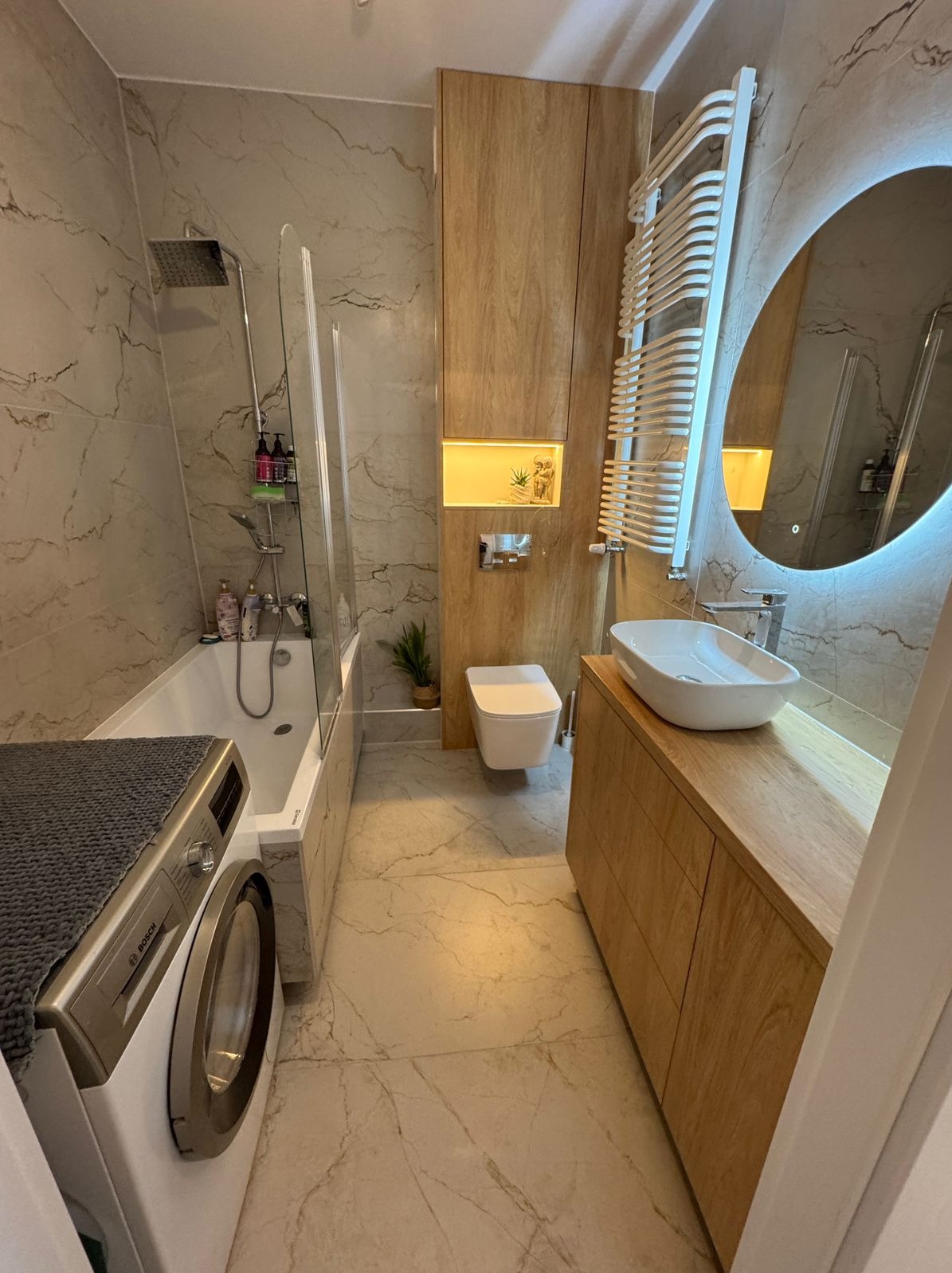 Nowoczesna łazienka z marmurowymi płytkami, drewnianą zabudową umywalki, owalnym lustrem z podświetleniem LED i pralką z szarym kocem na wierzchu.