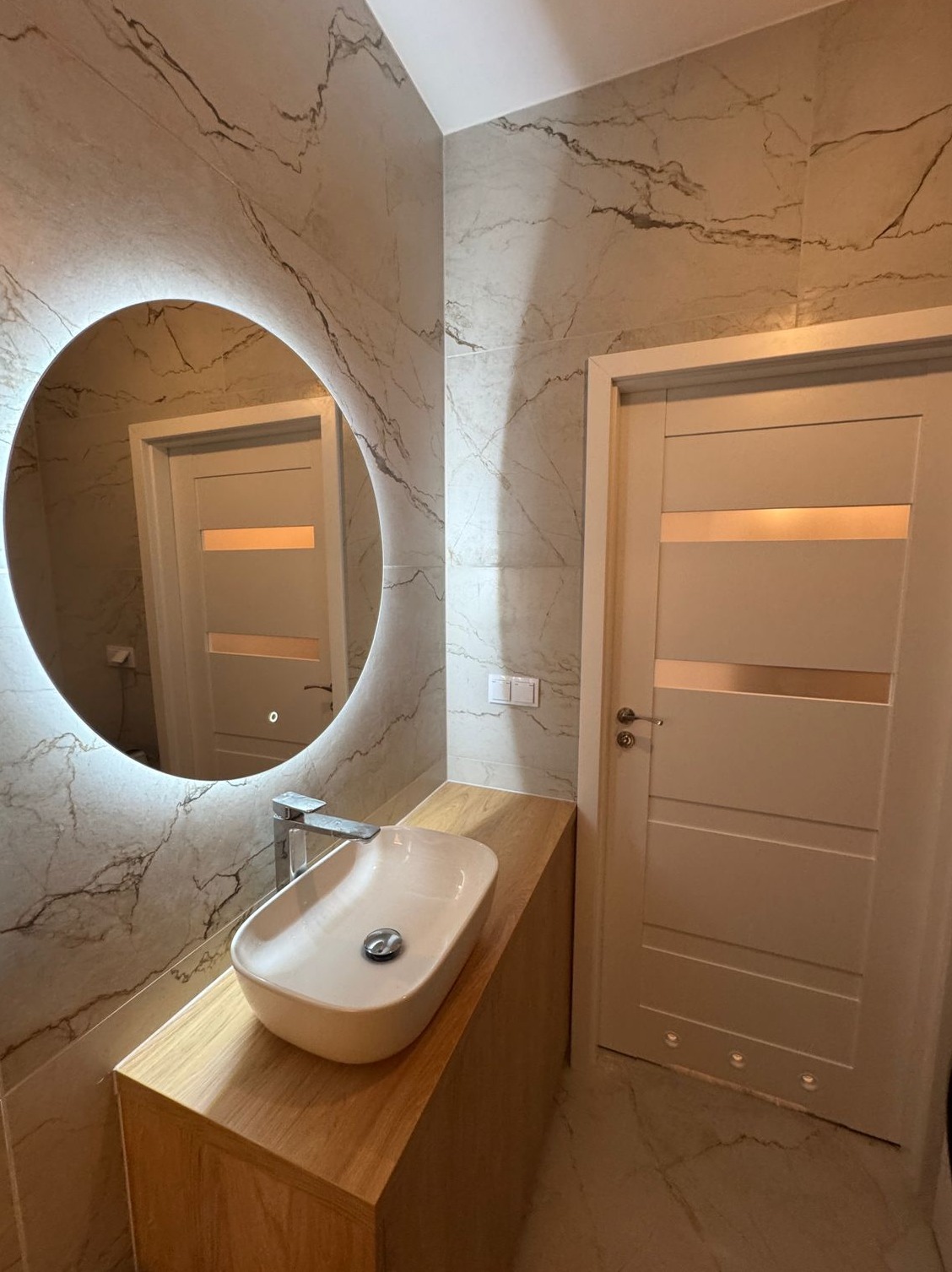 Nowoczesna łazienka z owalnym lustrem podświetlanym LED, umywalką nablatową na drewnianej szafce i drzwiami z poziomym przeszkleniem, wykończona płytkami imitującymi marmur.