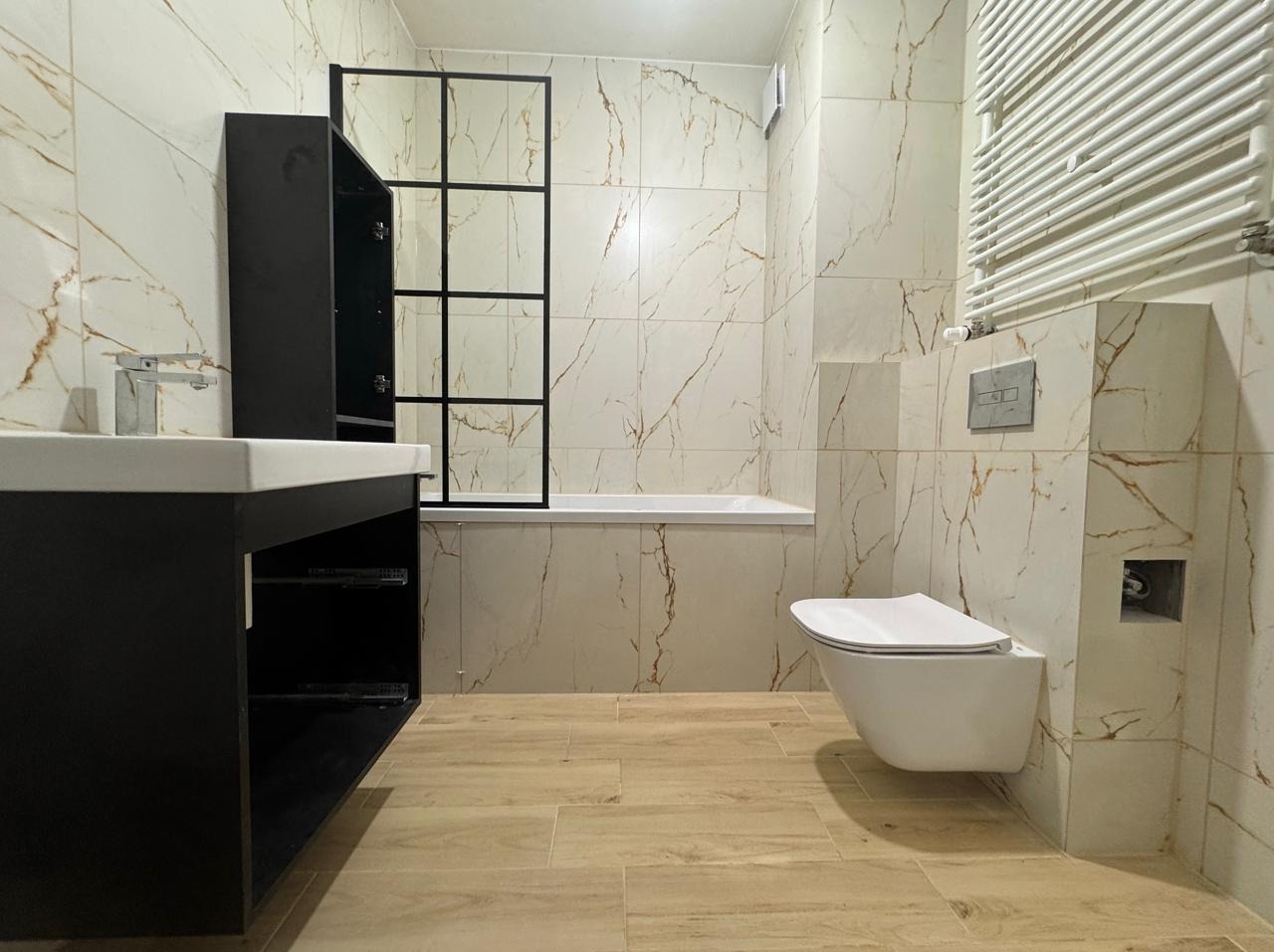 Wykończona łazienka w jasnych płytkach imitujących marmur z akcentami złota, z czarną szafką pod umywalką i kabiną prysznicową typu loft z czarnymi profilami, toaleta podwieszana.