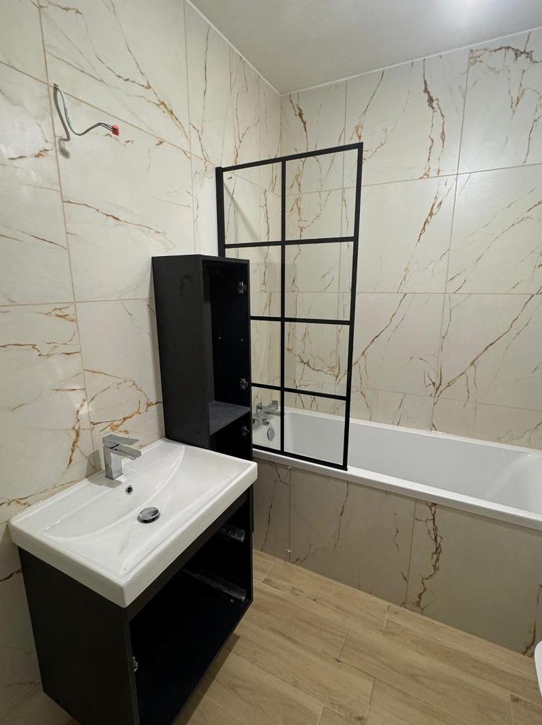 Nowoczesna łazienka z marmurowymi płytkami, czarną kabiną nawannową z geometrycznymi podziałami, białą umywalką z szafką w kolorze antracytu oraz drewnopodobną podłogą. Widoczne niedokończone...