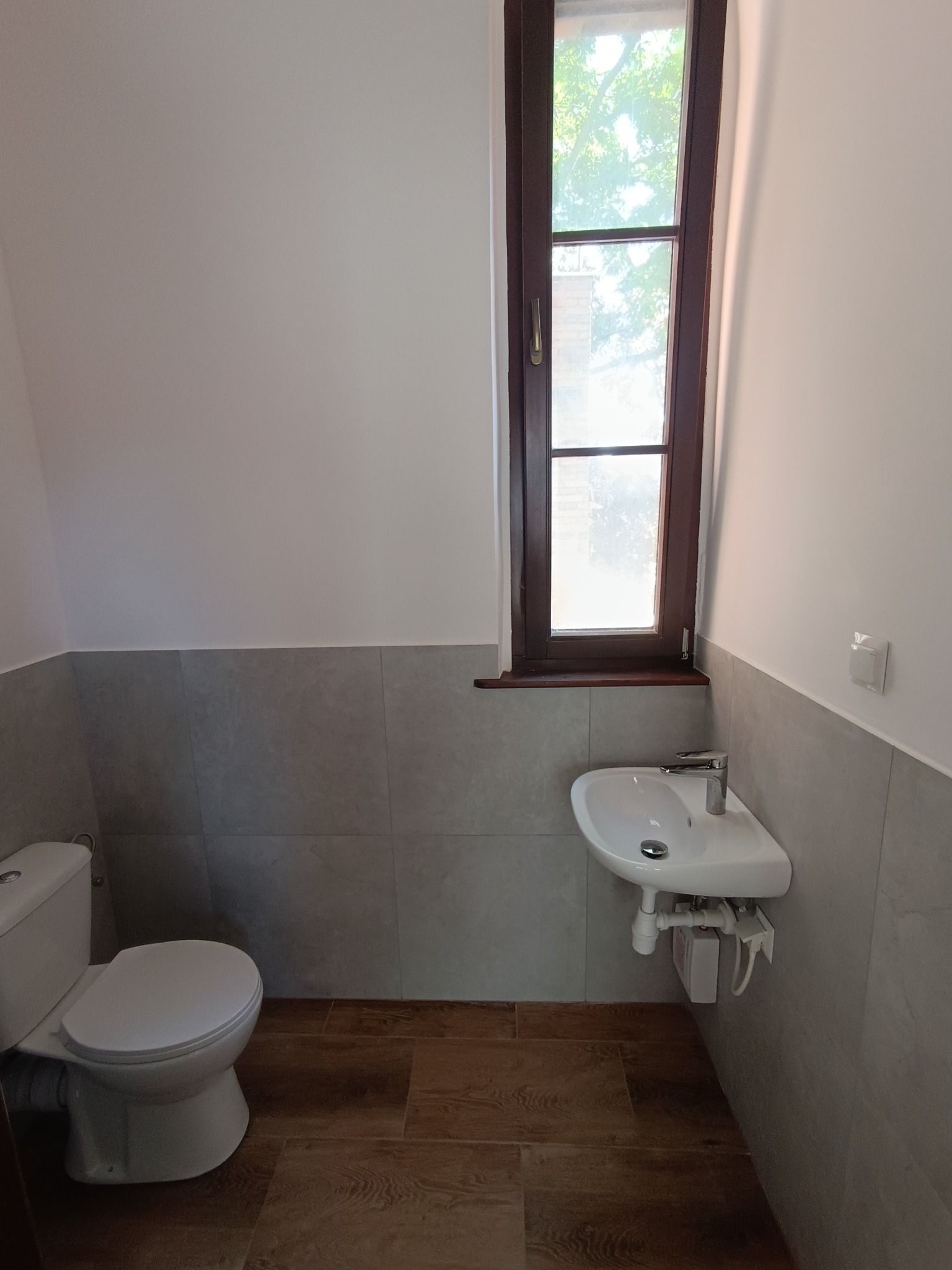 Jasna łazienka po remoncie: WC, umywalka pod oknem z brązową ramą, szare płytki do połowy ściany, drewnopodobna podłoga. Nowoczesne i funkcjonalne wykończenie.