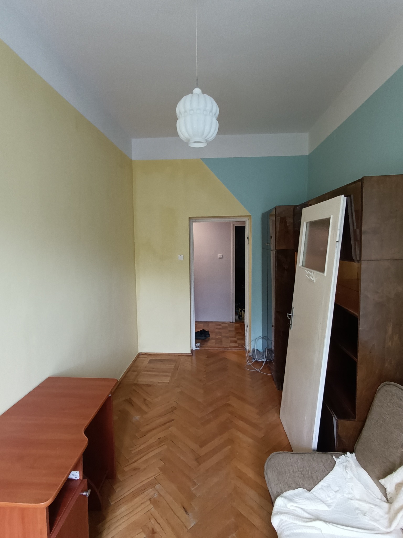 Pokój przed remontem: parkiet w jodełkę, ściany żółto-turkusowe z białym sufitem, wisząca lampa, meble w tle, drzwi oparta o szafę. Widok na wnętrze przez otwarte drzwi.