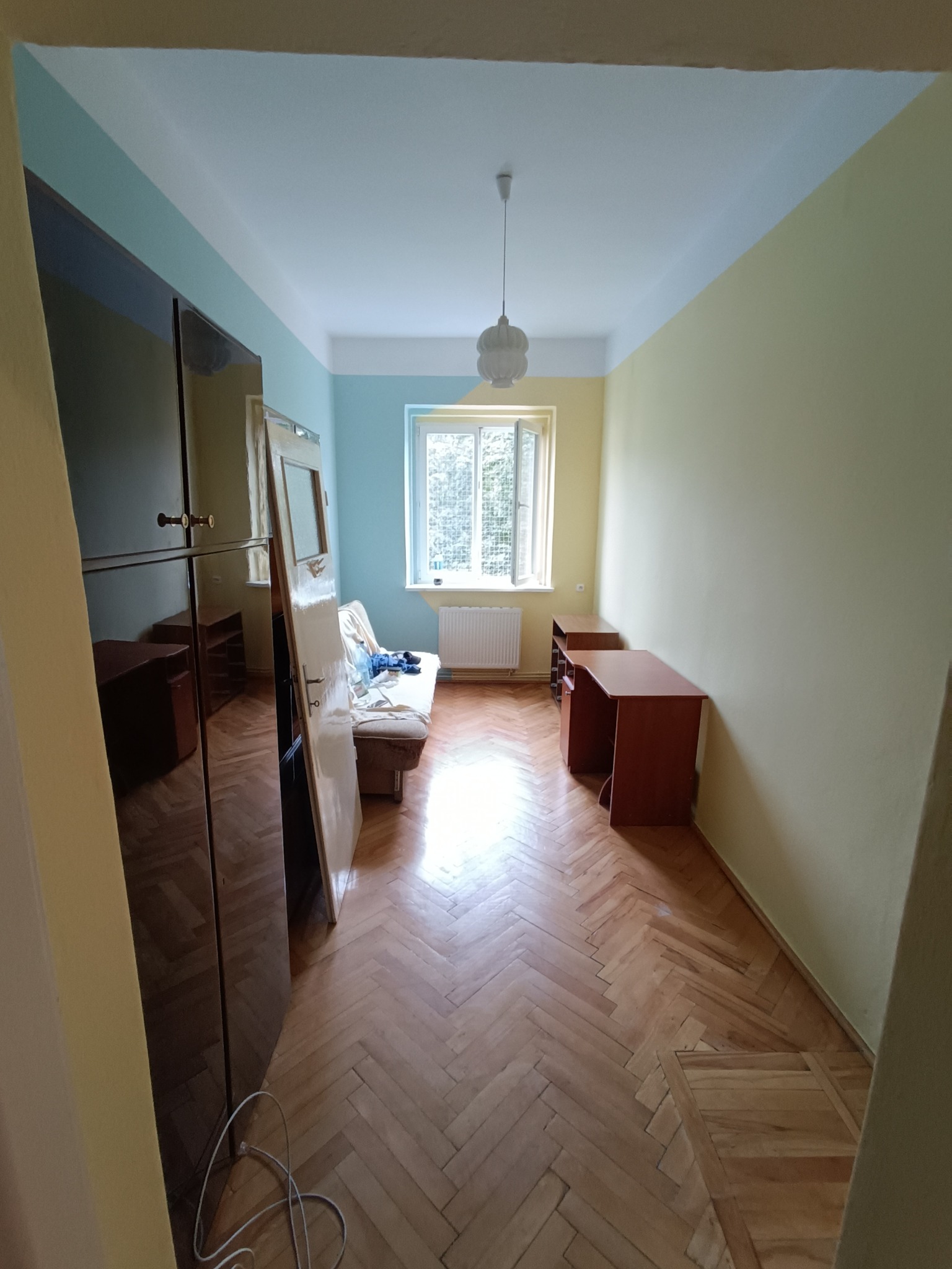 Pomalowane ściany na dwa kolory w pokoju z parkietem, widoczne meble i okno z grzejnikiem. Górna część ścian pomalowana na niebiesko, dolna na żółto.