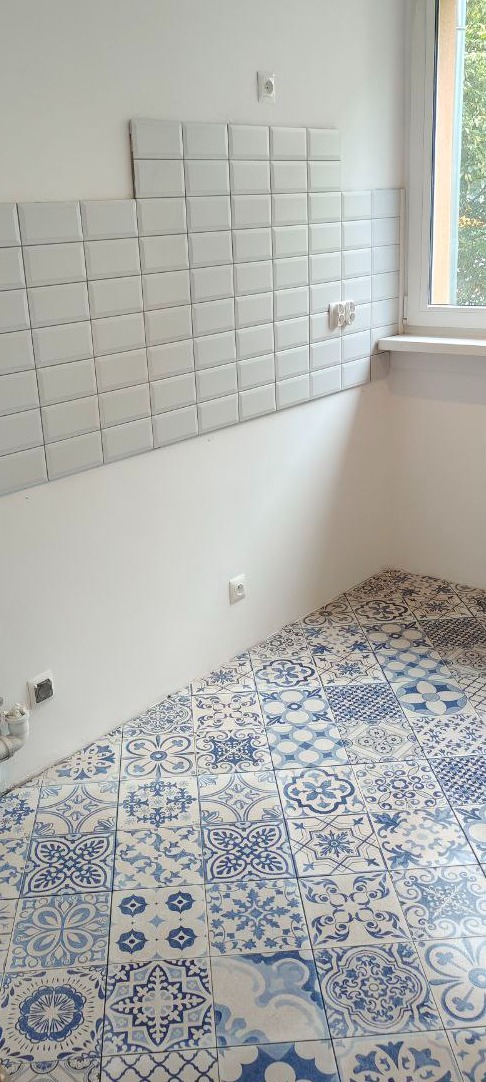 Ściana z białych płytek w stylu metro i podłoga z dekoracyjnych, niebiesko-białych płytek w rogu jasnego pomieszczenia z oknem. Częściowo ułożone płytki na ścianie.