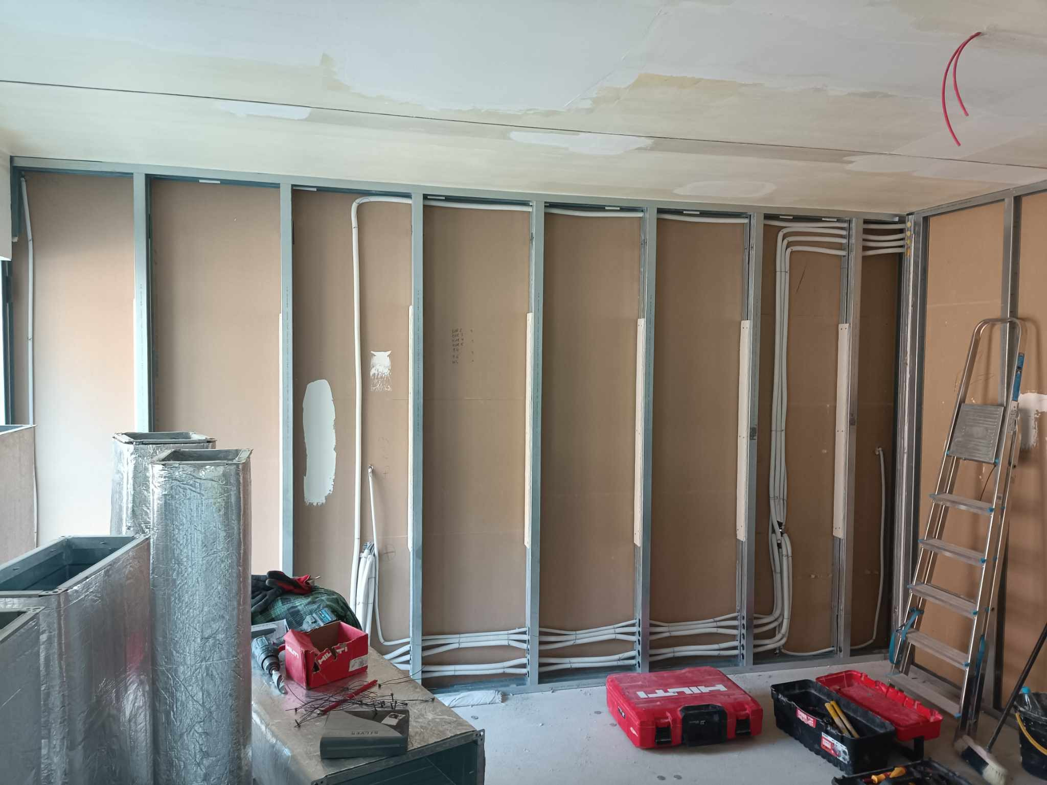 Montaż instalacji elektrycznej w ścianie z płyt karton gips: widoczne metalowe profile, przewody elektryczne w peszlach oraz częściowo zamontowane płyty. Na podłodze narzędzia i skrzynki narzędziowe.