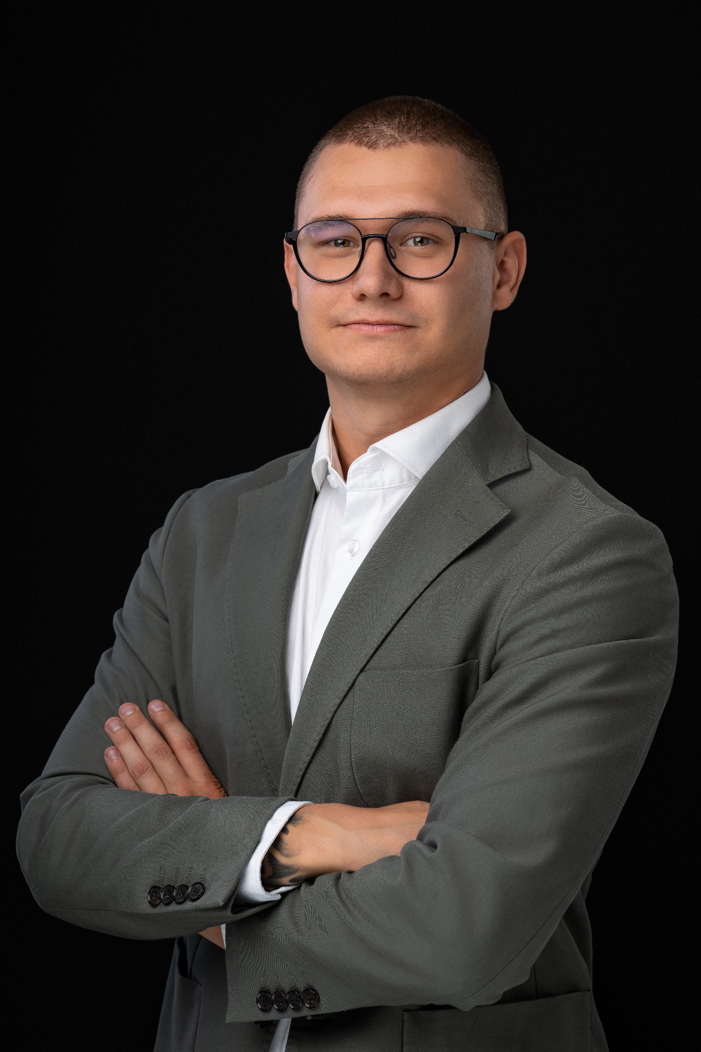 Portret mężczyzny w okularach i szarym garniturze na czarnym tle, z widocznym tatuażem na przedramieniu.