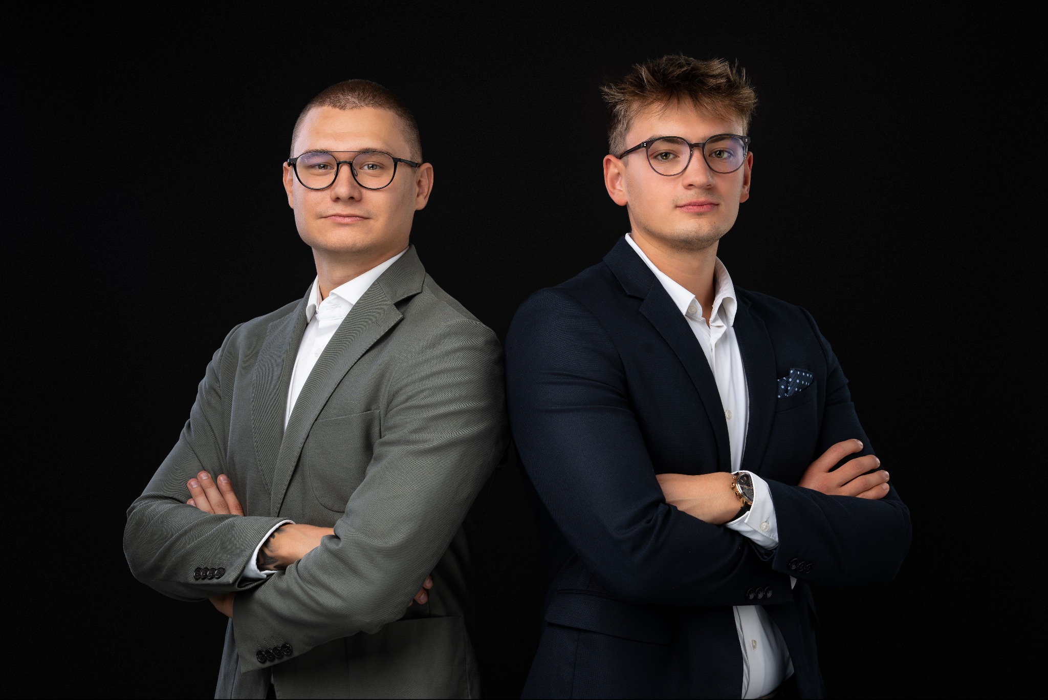 Portret dwóch młodych mężczyzn w garniturach, stojących plecami do siebie na czarnym tle, z rękami skrzyżowanymi na piersiach, obaj w okularach.
